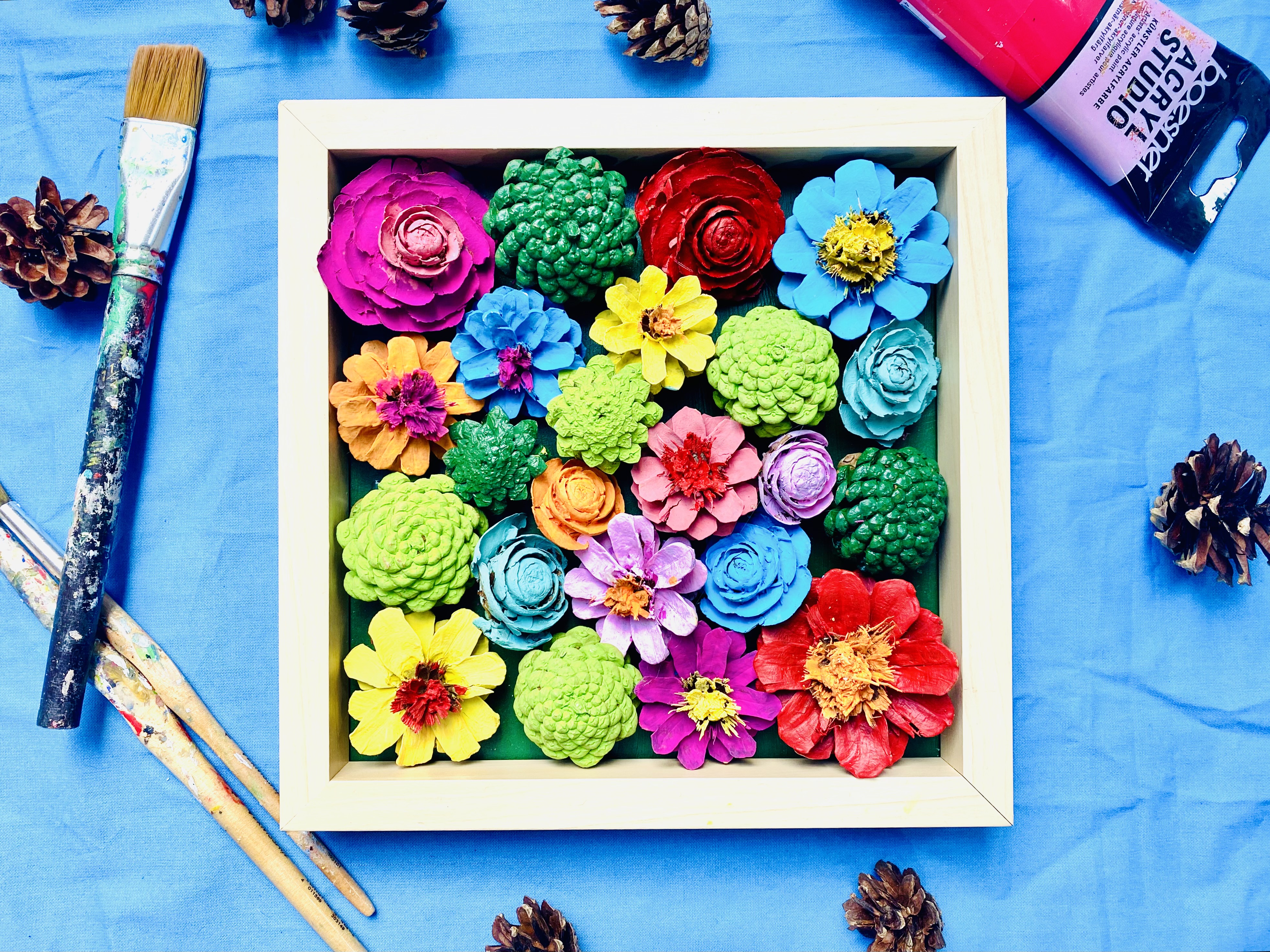 Das Bild zeigt einen Holzrahmen mit bunten Kiefernzapfen.