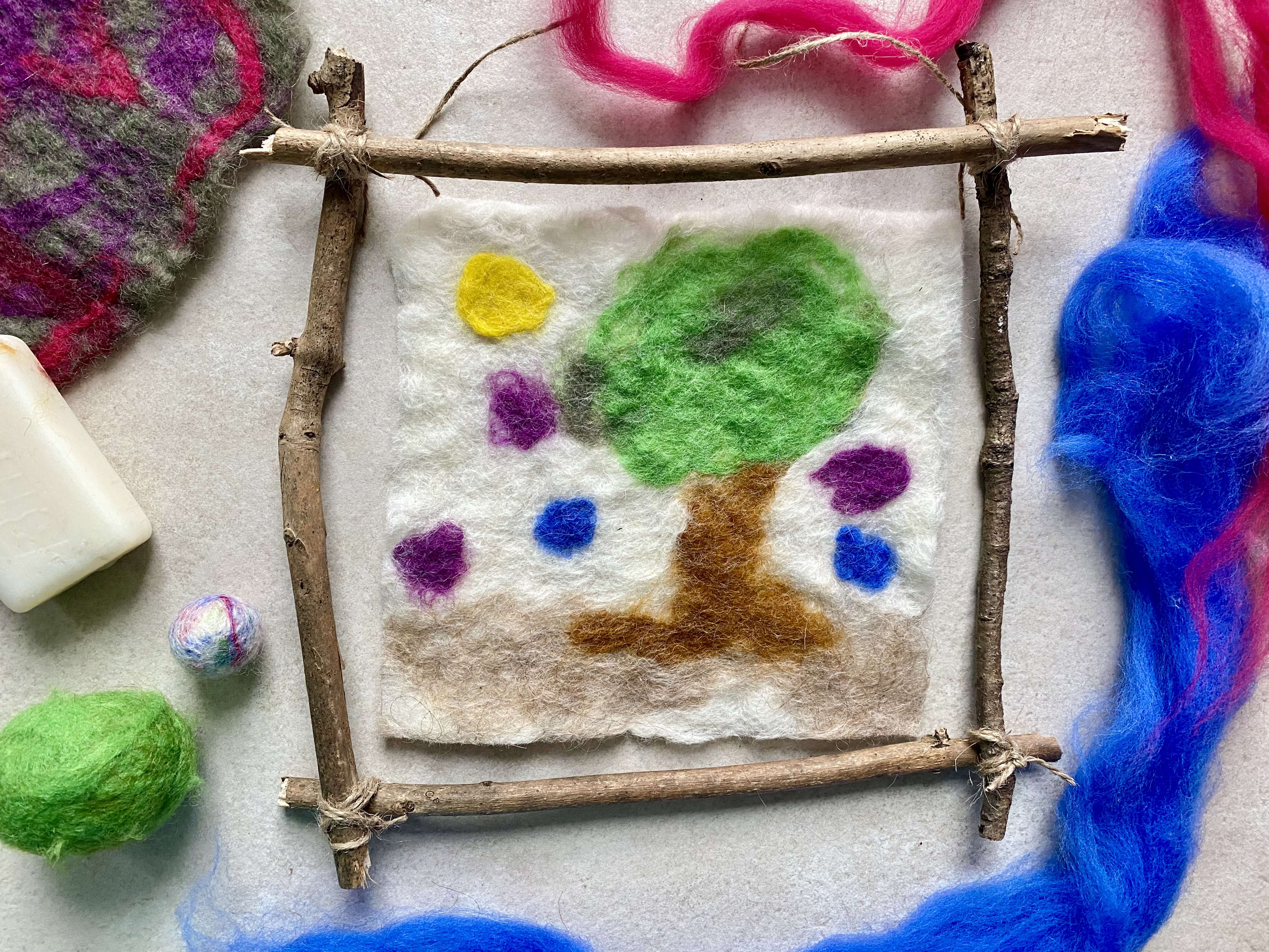Auf dem Bild ist ein Bilderrahmen aus Stöcken und einem aus Filz hergestellten Bild mit einem Baum zu sehen. Seife, bunte Kugeln und Filzwolle liegen um das Bild herum. 
