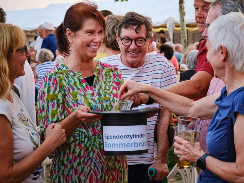 Ein Mensch hält einen schwarzen Hut falsch herum. Andere Menschen werfen Geld in den Hut hinein. 