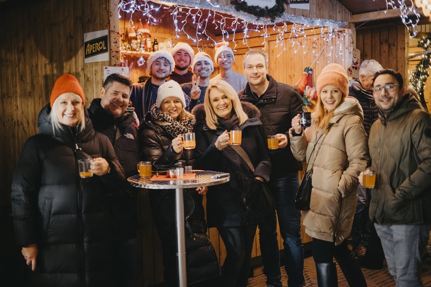 Das Bild wurde auf dem Friedrichsdorfer Weihnachtsmarkt aufgenommen. Eine Gruppe Menschen lächelt in die Kamera. Alle sind winterlich angezogen und halten eine Tasse mit einem Heißgetränk in der Hand. 