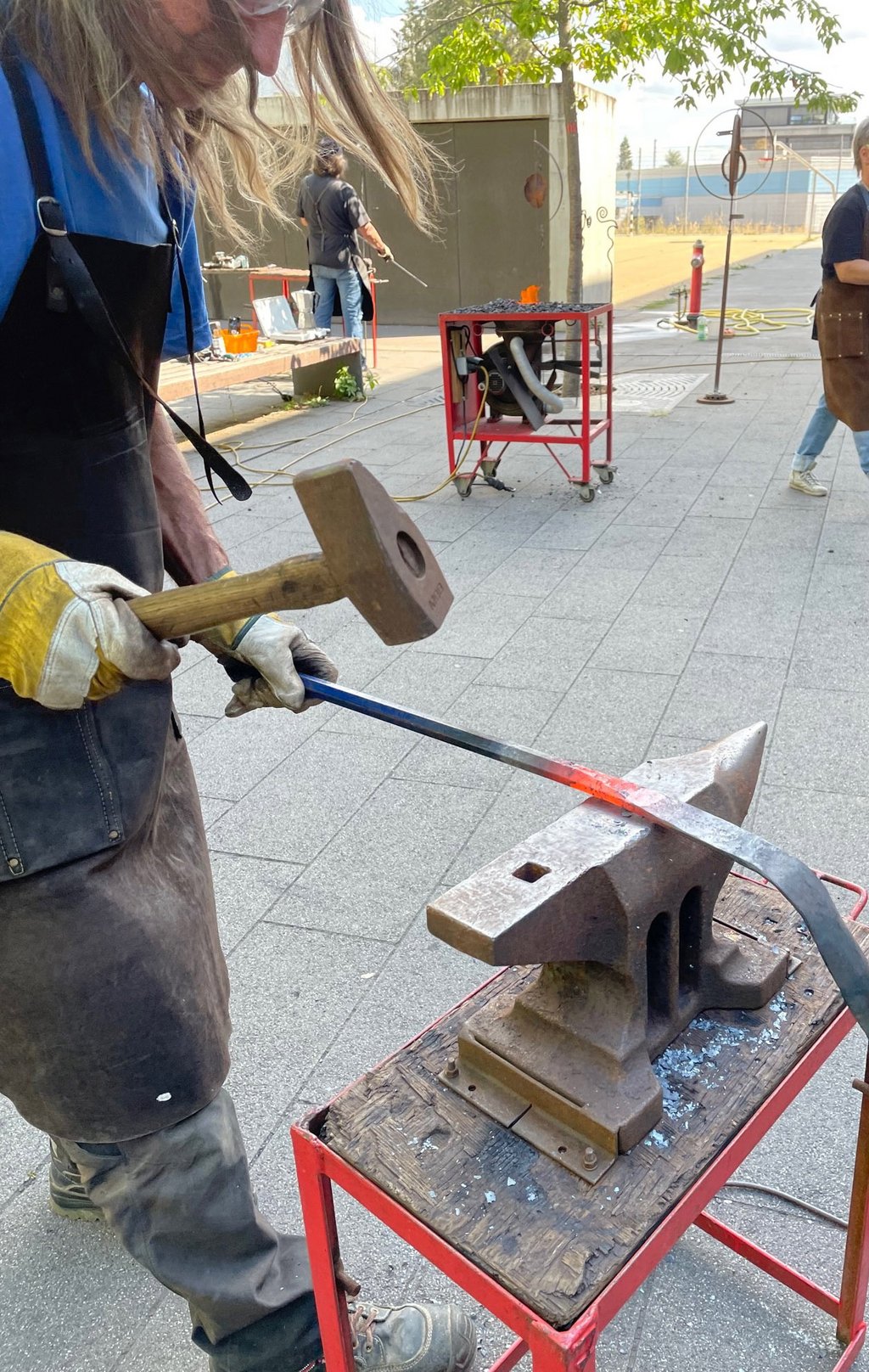 Auf dem Bild ist ein Mann zu sehen, der ein glühendes Stück Eisen mit einem Hammer bearbeitet.