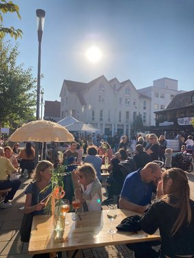 Viele Personen mit Getränken auf den Tischen in entspannter Atmosphäre bei Sonnenschein auf einem Platz in einer Innenstadt. 