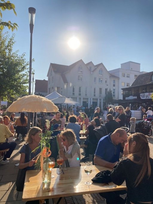 Viele Personen mit Getränken auf den Tischen in entspannter Atmosphäre bei Sonnenschein auf einem Platz in einer Innenstadt. 