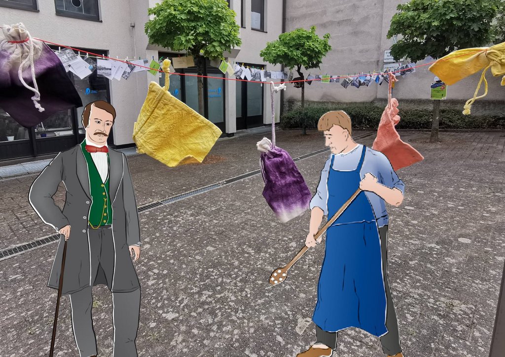 Zwei stilisierte Figuren stehen im Freien auf einem gepflasterten Platz vor einem Gebäude mit großen Fenstern und Bäumen im Hintergrund. Links steht eine Person in einem grauen Anzug mit grüner Weste, einem roten Halstuch und einem Stock in der Hand. Rechts steht eine Person in einem blauen Overall, die einen großen Holzlöffel hält. Über den Figuren hängen an einer Leine bunter Säckchen, die an Wäscheklammern befestigt sind. Im Hintergrund sind weitere Bilder an der Leine zu sehen.