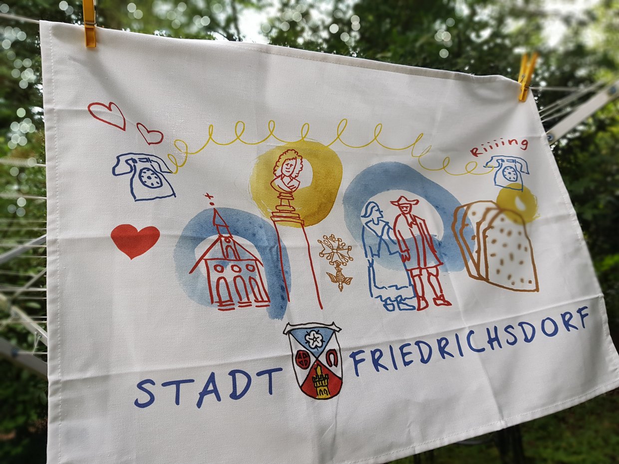 Auf dem Bild ist ein weißes Geschirrhandtuch mit einer bunten Comiczeichung zur Friedrichsdorfer Stadtgeschichte darauf zu sehen.