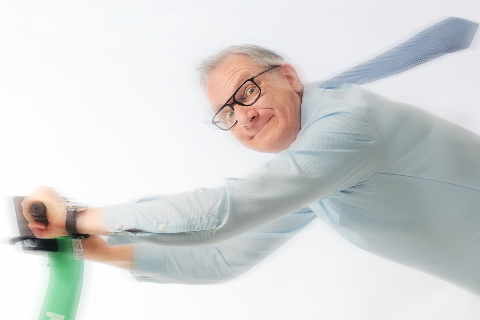 Auf dem Bild ist Hans Gerzlich mit fliegender Krawatte zu sehen, der sich am Lenker eines grünen E-Rollers festhält.