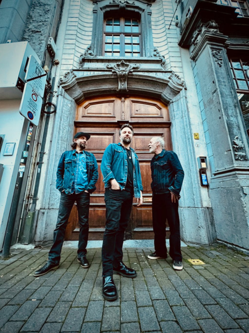 Auf dem Bild sind die drei Musiker vom Gerry Hundt Trio vor einem großen hölzernen Portal stehend zu sehen.