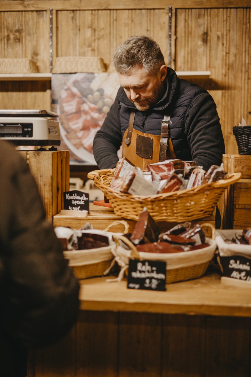 Auf dem Bild sieht man einen Ausschnitt eines Standes auf dem Weihnachtsmarkt. Auf der Verkaufsfläche sind Körbe mit Schinken gefüllt. Der Verkäufer steht hinter dem Stand und blickt nach unten. 