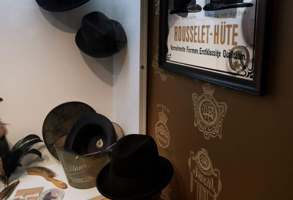 Das Bild zeigt eine Ausstellungsvitrine mit mehreren schwarzen Hüten, darunter ein Zylinder, der falschherum in einer Schachtel liegt. In seinem Inneren ist ein etikett zu sehen. Im Hintergrund hängt ein gerahmter Spiegel mit der Aufschrift "ROUSSELET-HÜTE - Vornehmste Formen, Erstklassige Qualitäten." Am der Wand sind verschiedene Logos und Schriftzüge von Hutfabriken zu sehen.