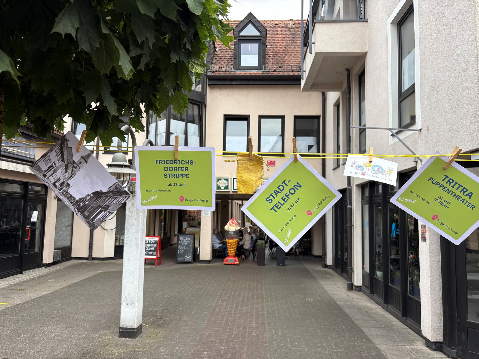Back to Gängelsche - Philipp-Reis-Passage Das Bild zeigt einen Ausschnitt einer gespannten Wäscheleine, an der verschiedene Zettel mit Veranstaltungshinweisen befestigt sind. Im Hintergrund ist die Philipp-Reis-Passage zu erkennen.