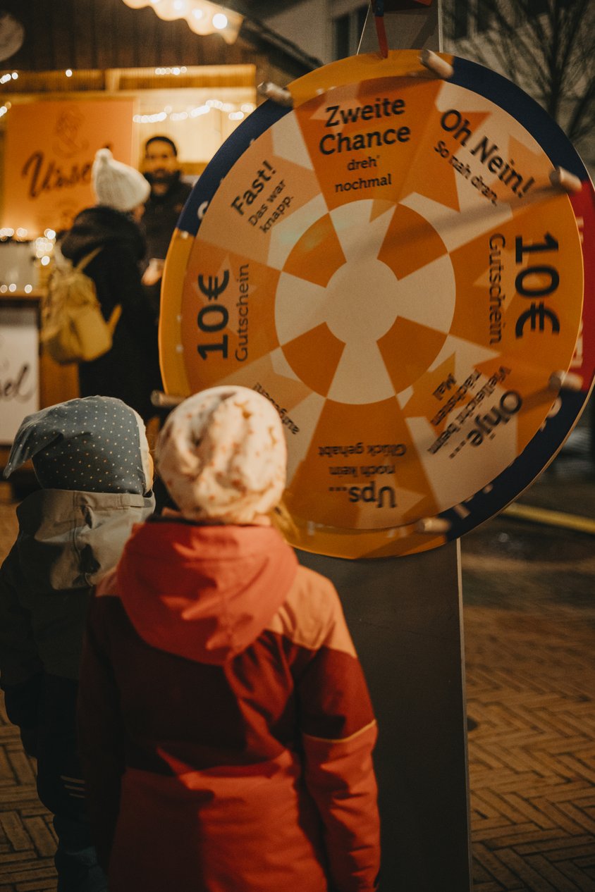 Auf dem Bild sieht man zwei Kinder mit dem Rücken zum Betrachtenden. Die Kinder sind winterlich angezogen und stehen vor einem Glücksrad. Das Sie scheinbar gerade angedreht haben. 