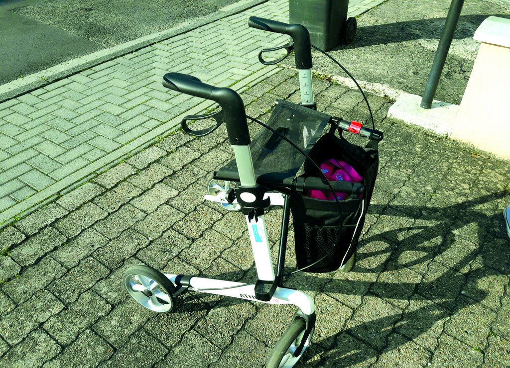 ein faltbarerer Rollator auf dem Bürgersteig mit festgestellter Bremse und pinkem Inhalt im Korb