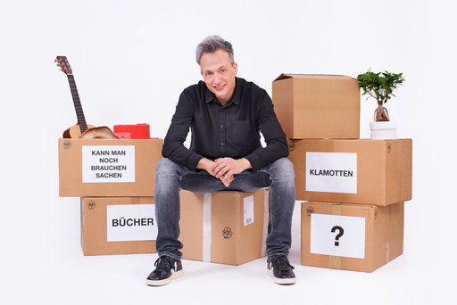Auf dem Bild ist Olaf Bossi zu sehen, der auf einem Umzugkarton sitzt.