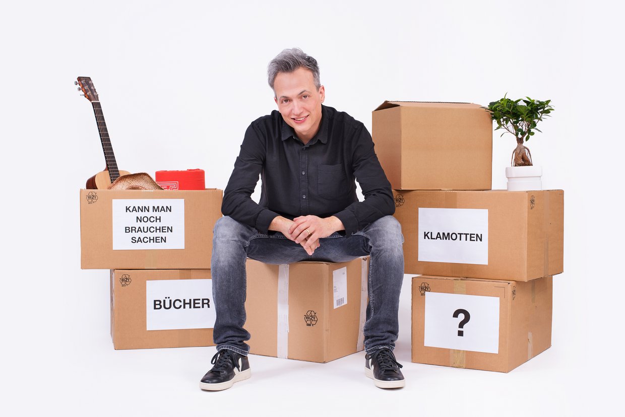 Auf dem Bild ist Olaf Bossi zu sehen, der auf einem Umzugkarton sitzt.