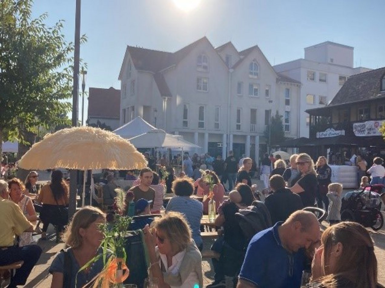 Viele Personen mit Getränken auf den Tischen in entspannter Atmosphäre bei Sonnenschein auf einem Platz in einer Innenstadt.