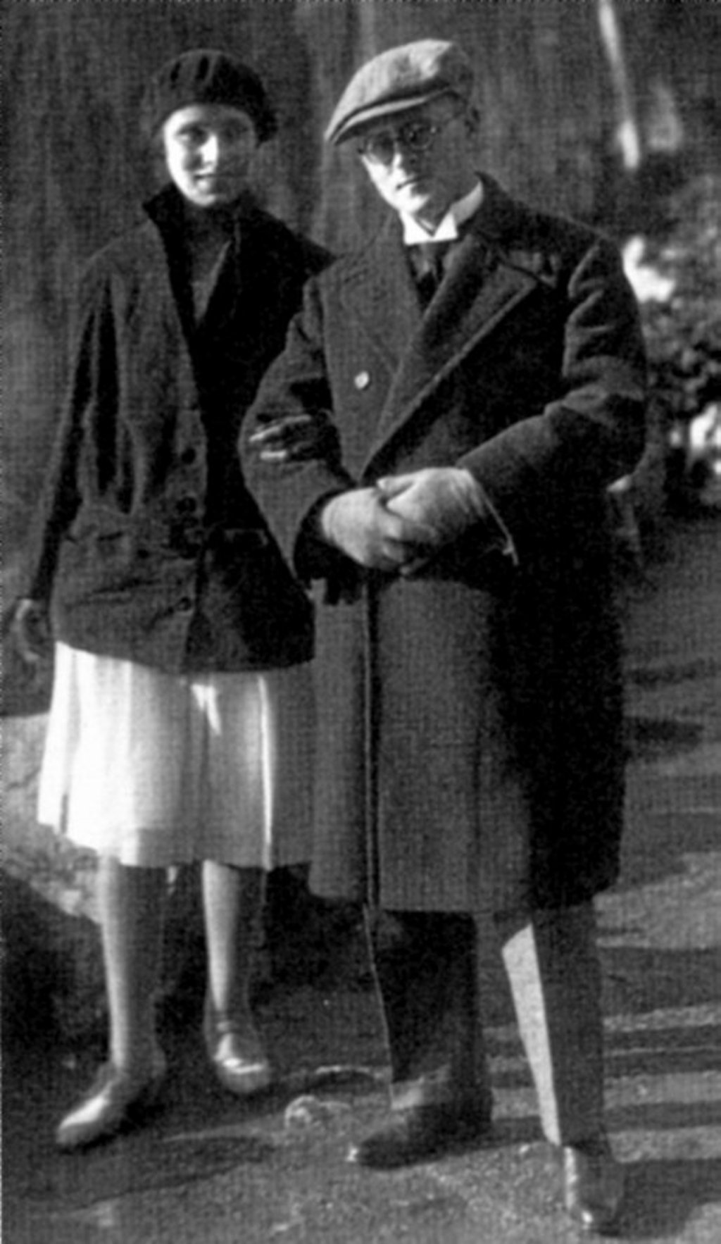 Schwarz-Weiß-Fotografie des Ehepaares Neiß im Freien. Ehefrau Elfriede im hellen Rock und dunkler Jacke steht neben ihrem Ehemann Dr. Friedrich Neiß mit Brille, Schiebermütze und langem Mantel. Beide blicken in die Kamera und stehen dicht beieinander auf einem sonnenbeschienenen Weg.