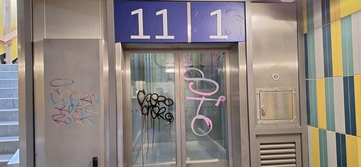 Auf dem Bild ist der Aufzug der Unterführung, welcher zu den Gleisen 1 und 11 führt, sichtbar. Die metallene Aufzugsanlage ist mit mehreren farbigen Graffiti-Tags beschmiert, darunter schwarze und rosafarbene Schriftzüge auf den Glastüren sowie ein buntes Graffiti auf der linken Metallwand.