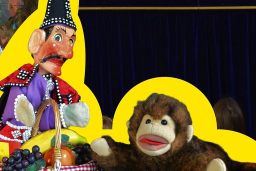 Eine bunte Puppentheaterbühne mit Vorhang und goldener Verzierung steht im Freien. Auf der Bühne sind zwei Puppen in traditioneller Kleidung zu sehen. Vor der Bühne steht links eine große Puppe mit spitzem Hut und violettem Gewand, die einen Korb mit Obst hält. Rechts davon sitzt ein braunes Stofftier in Form eines Affen mit rotem Mund und ausladenden Armen. Im Hintergrund sind bunte Wimpel und ein Baum zu erkennen. Vor der Bühne sitzen Kinder und Erwachsene als Publikum.