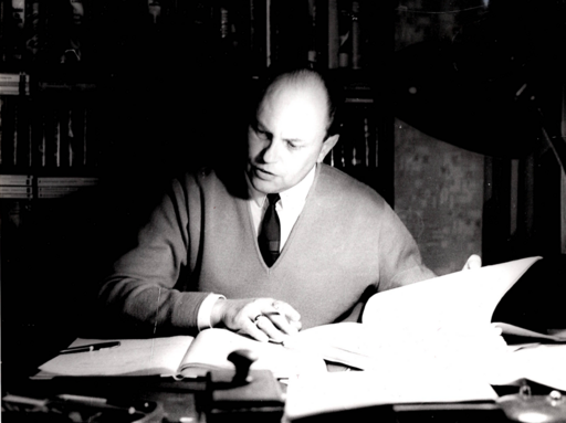 Karl-Herbert Scheer sitzt an seinem Schreibtisch und betrachtet ein Buch sowie darum verstreute Unterlagen. Er trägt ein weißes Hemd, darüber einen Pullover und eine Krawatte. Im Hintergrund stehen Bücherregale.