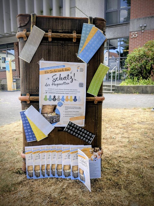 Ein dekorierter historischer Koffer mit bunten Stickern und Flyern zur dritten Friedrichsdorfer Stadtrallye steht vor dem Rathaus. Die Materialien erinnern an den erfolgreichen Abschluss der dritte Friedrichsdorfer Schatzsuche.