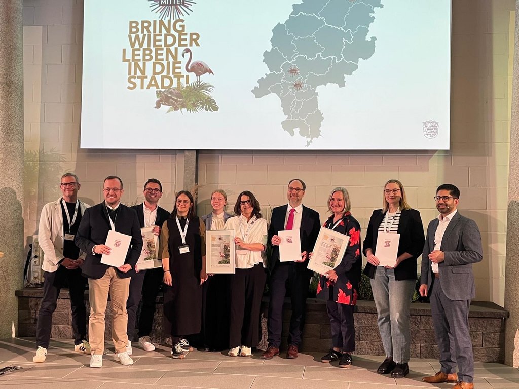 Auf dem Bild sind die Preisträgerinnen und Preisträger des Innenstadtwettbewerbs „Ab in die Mitte“ aus den Städten Friedrichsdorf, Frankfurt und Melsungen gemeinsam auf der Bühne mit Kaweh Mansoori zu sehen. Im Hintergrund ist das diesjährige Motto des Wettbewerbs „Bring wieder Leben in die Stadt“ abgebildet. 