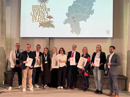 Auf dem Bild sind die Preisträgerinnen und Preisträger des Innenstadtwettbewerbs „Ab in die Mitte“ aus den Städten Friedrichsdorf, Frankfurt und Melsungen gemeinsam auf der Bühne mit Kaweh Mansoori zu sehen. Im Hintergrund ist das diesjährige Motto des Wettbewerbs „Bring wieder Leben in die Stadt“ abgebildet. 