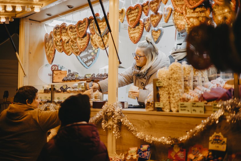 Das Bild zeigt einen Stand auf dem Friedrichsdorfer Weihnachtsmarkt. Im Vordergrund sind zwei Personen abgebildet die mit dem Rücken zum Betrachtenden gerade etwas an einem Süßwarenstand kaufen. Der Stand ist üppig mit Lebkuchenherzen und anderen Süßigkeiten dekoriert. 