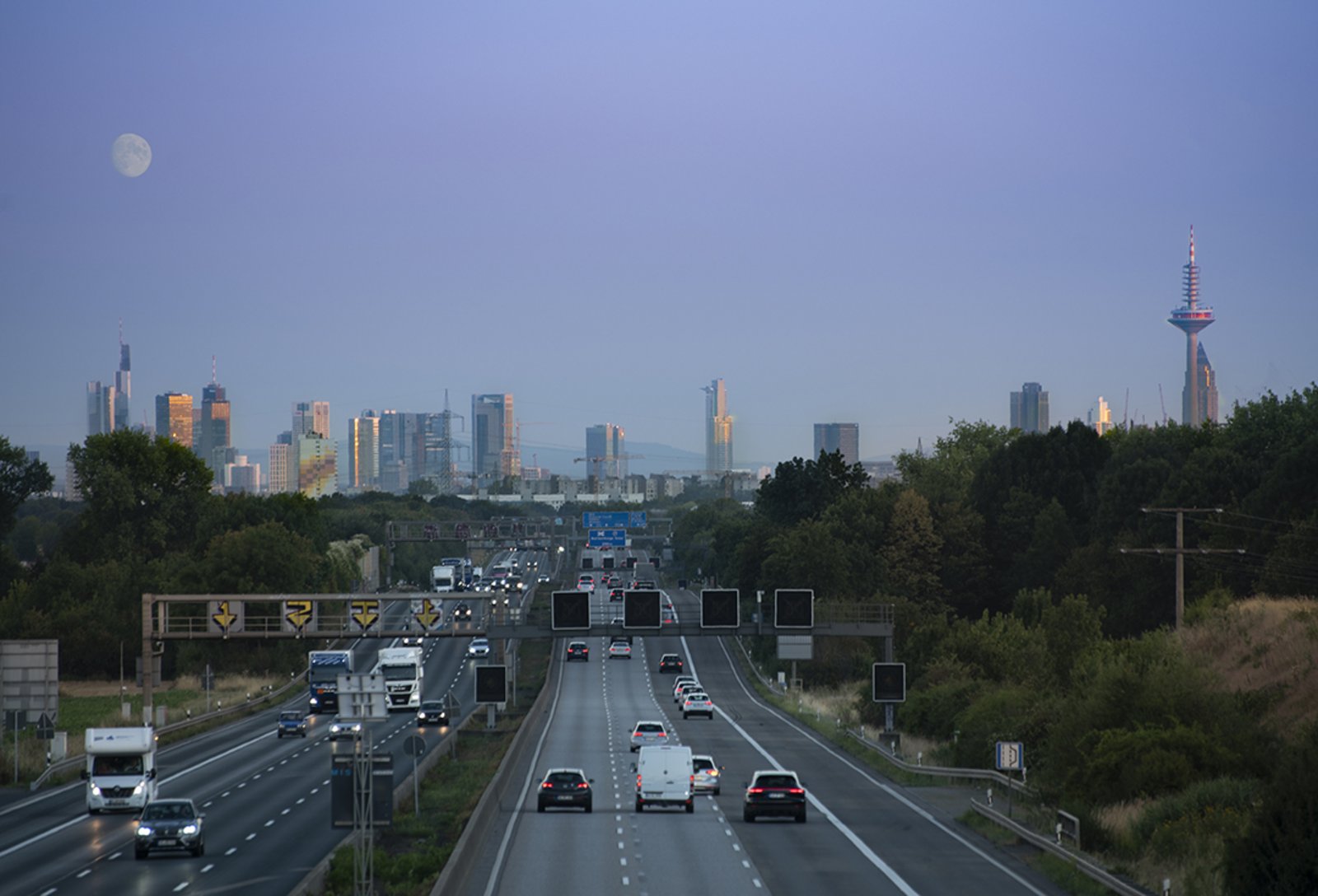 Zu sehen sind die Autobahn BAB 5, die Skyline Frankfurts und der Mond in Abenddämmerung aus der Perspektive Friedrichsdorf. 