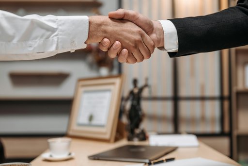 Zwei Personen reichen sich in einem Büro die Hand. Eine Person trägt ein weißes Hemd, die andere einen schwarzen Anzug. Auf dem Schreibtisch im Hintergrund stehen eine Tasse, ein Laptop, Schreibutensilien sowie ein Bilderrahmen. Außerdem ist eine Justitia-Figur sichtbar, was auf ein juristisches Umfeld hinweist.