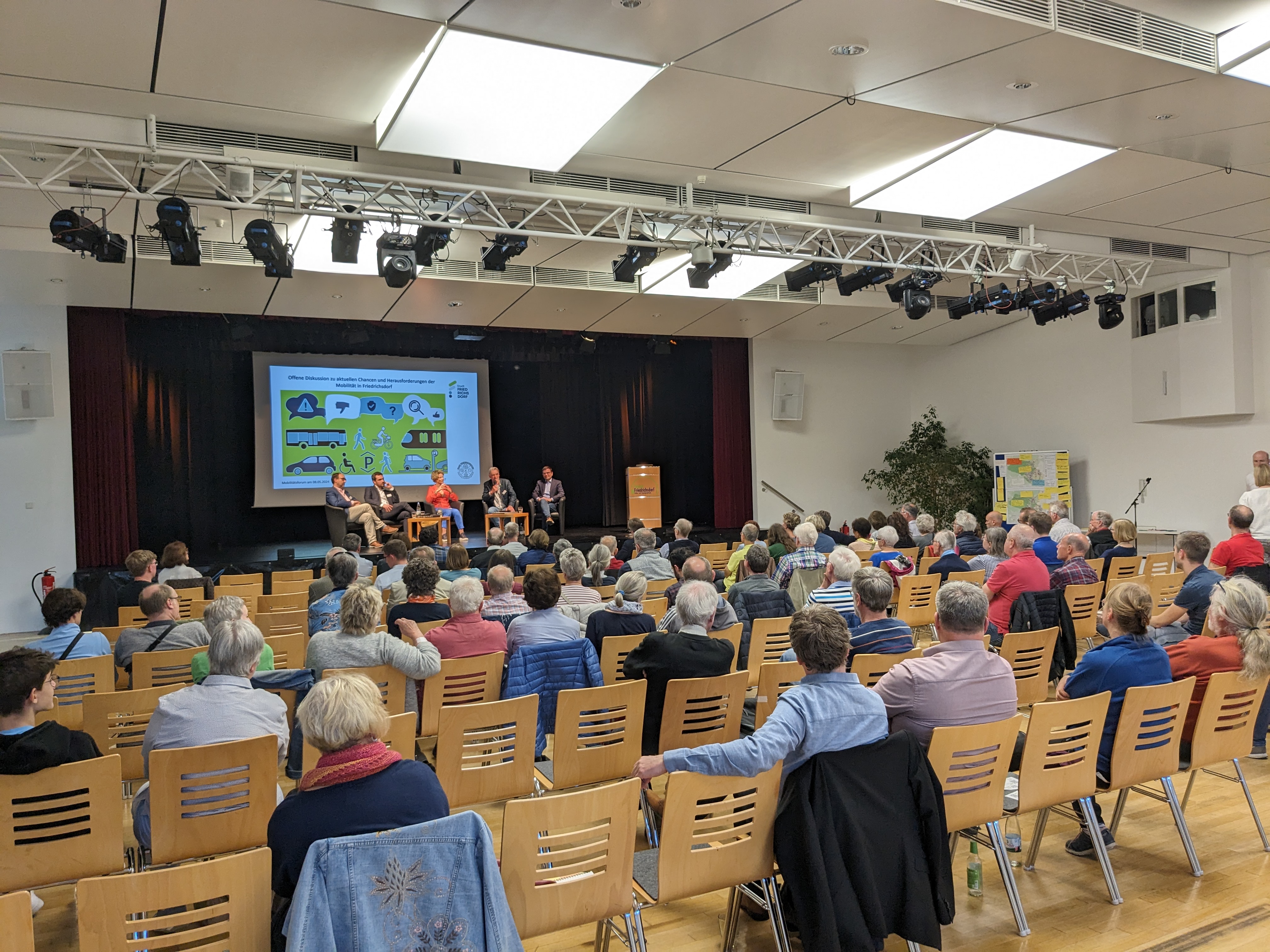 Zeigt Personen im Forum Friedrichsdorf, welche einem Vortrag auf der Bühne folgen.