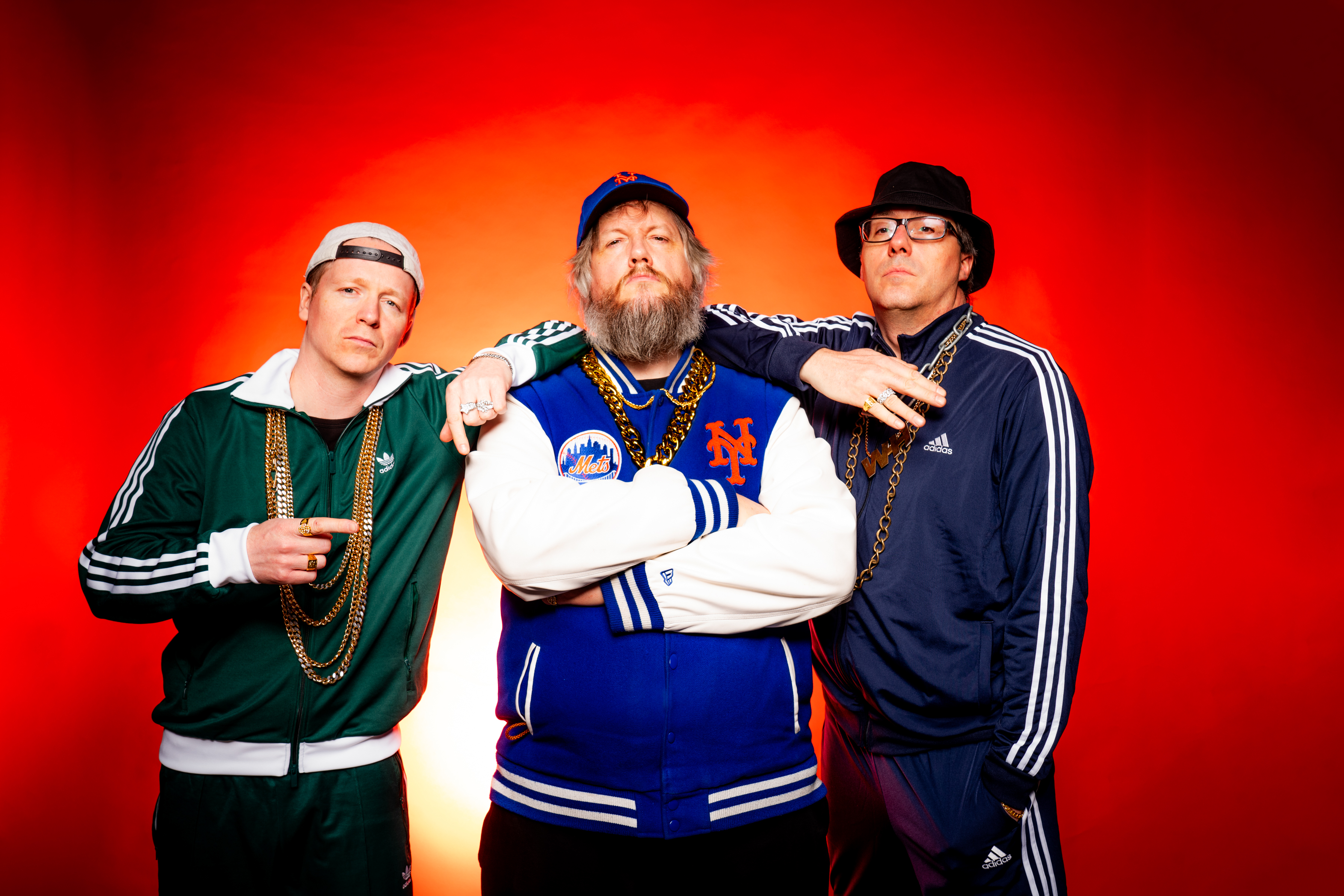 Auf dem Bild ist das Trio Wildes Holz in bunten Trainigsanzügen mit Goldketten vor rotem Hintergrund zu sehen.