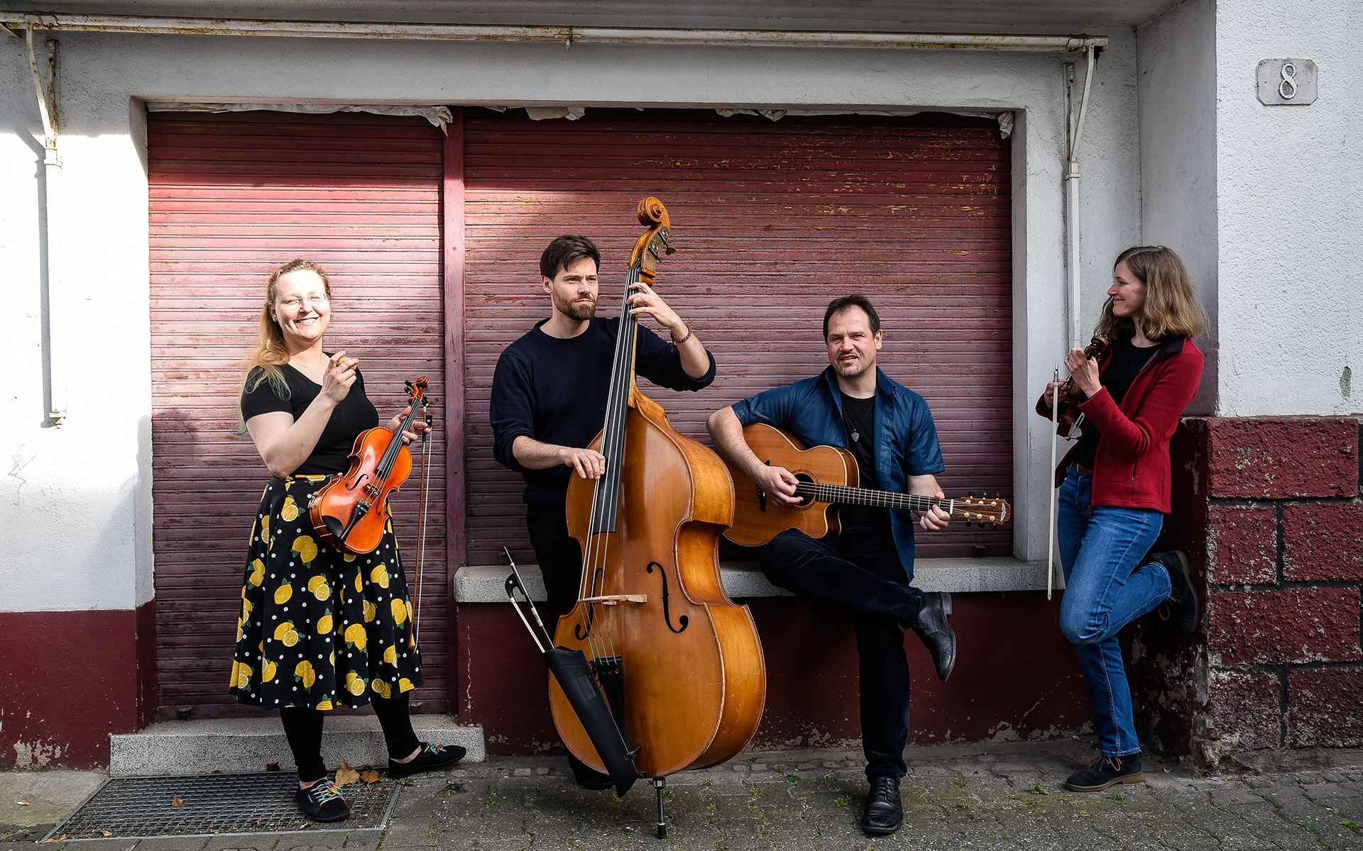 Auf dem Bild sind die vier Musiker der Band Feinschmeckerfolk mit ihren Instrumenten vor einem Rolladen stehend zu sehen.