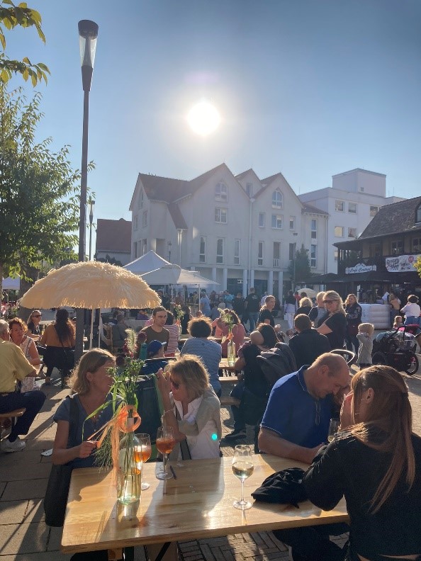 Viele Personen mit Getränken auf den Tischen in entspannter Atmosphäre bei Sonnenschein auf einem Platz in einer Innenstadt. 